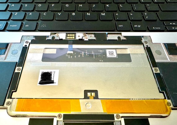 Underside of the Framework Laptop 16 trackpad module sitting on the keyboard deck of the laptop. There are several layers of non-conductive tape placed on either side of the actuation mechanism indentation of the support bar for the trackpad as an attempt to reduce the play in the trackpad surface during normal use.